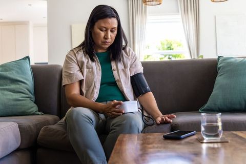 Asian woman checking blood pressure at home with digital cuff, smartphone, glass of water