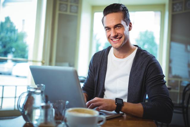 Man smiling while using laptop in a cozy café, with a coffee cup in the foreground. Ideal for illustrating themes of remote work, freelance lifestyle, productivity, and modern casual work environments. Suitable for blogs, articles, or websites focusing on technology, work-life balance, and digital nomadism.
