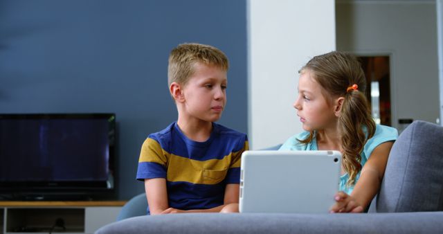Children sitting together on sofa using a digital tablet. Young boy in striped shirt and girl in blue shirt. Ideal for themes on modern family life, technology education, and indoor activities.