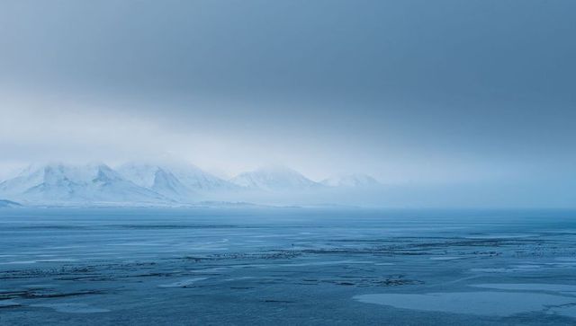 Arctic coast revealing frozen sea, misty snow-capped peaks, icy leads and minimalist blue seascape