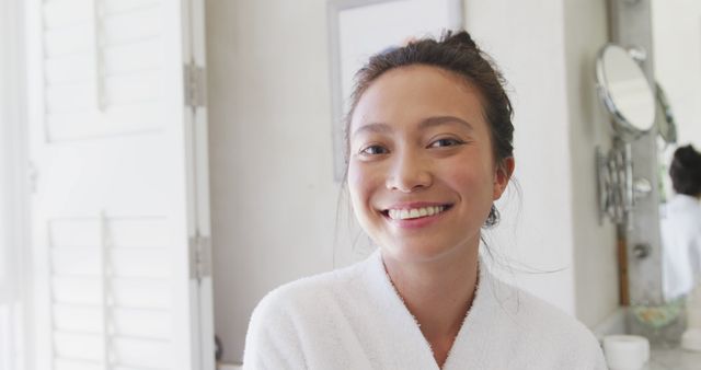 Smiling woman wearing white robe standing in a clean and minimalist bathroom filled with natural light. Ideal for promoting morning routines, skincare products, health and wellness initiatives, and self-care campaigns. The serene and calm atmosphere lends itself well to advertisements for home accessories, personal hygiene products, and spa services.
