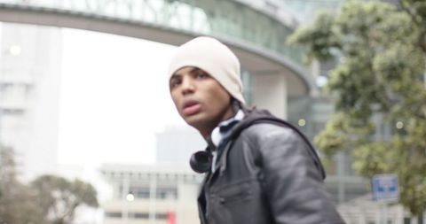 Young man standing on urban sidewalk wearing beanie and headphones under pedestrian bridge