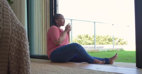 African american woman sitting at sliding glass door holding mug enjoying sunlit patio