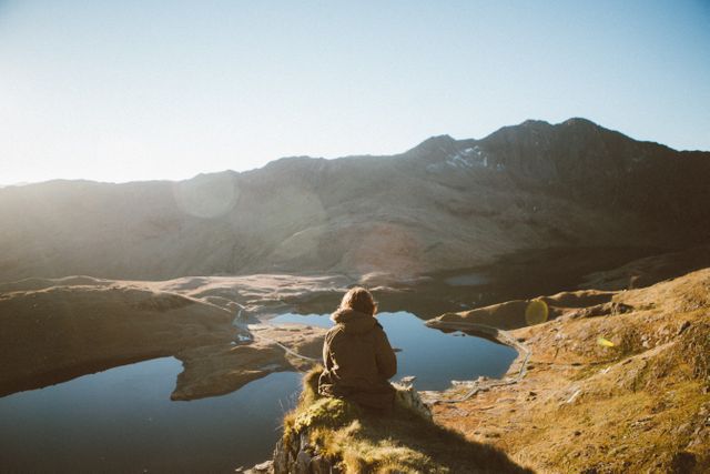 Lone hiker is sitting on a grassy cliff edge, admiring the calm lake nestled between rugged mountain peaks at sunrise. Great for use in travel blogs, adventure advertising, and mental health articles highlighting the benefits of solitude and nature.