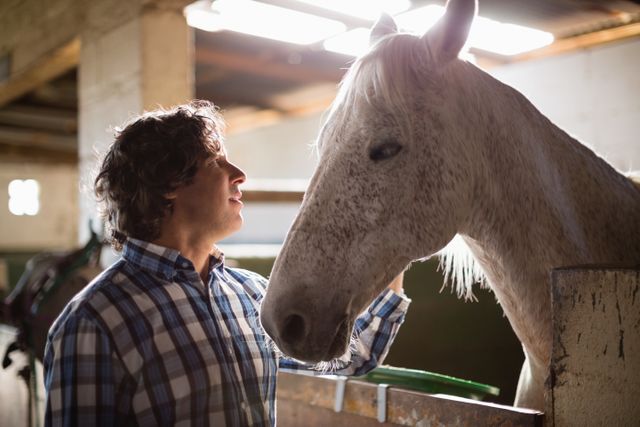 Man gently caressing a horse in a stable setting. The man is wearing a plaid shirt and smiling, showing a bond between human and animal. This image can be used for themes related to livestock care, animal companionship, farm life, equestrian activities, and the human-animal bond.