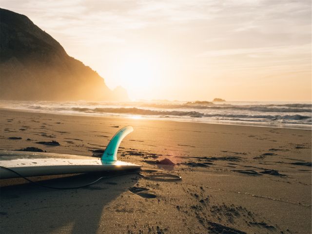 Surfboard lying on sandy beach with ocean waves and setting sun in background, near rocky coastline. Perfect for themes related to surfing, adventure travel, beach holidays, watersports, and coastal serenity.