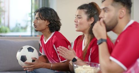 Friends in red jerseys cheering soccer match on couch holding ball and sharing popcorn
