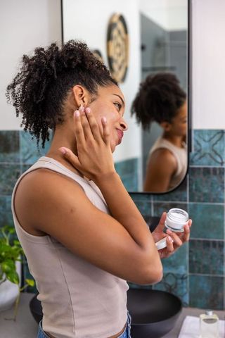 African american woman applying moisturizer to cheek at bathroom vanity mirror
