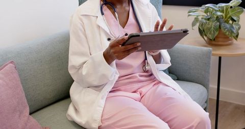 Clinician reviewing tablet chart on clinic sofa wearing white lab coat and pink scrubs