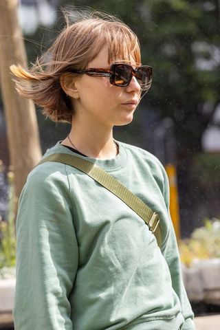 Young woman wearing tortoise sunglasses and green sweatshirt standing in sunny urban breeze