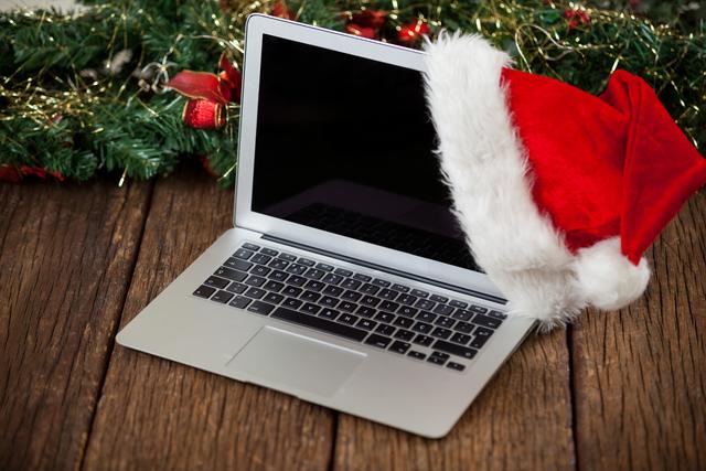 This image features a laptop with a Santa hat placed on a rustic wooden table, adorned with Christmas decorations including pine branches and gold garland. It captures the festive spirit and can be used for holiday-themed marketing, social media posts, and seasonal promotions, especially those related to technology and holiday sales.