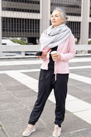 Confident senior woman standing on urban plaza holding coffee and smartphone wearing scarf
