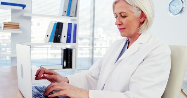 Senior female doctor wearing white coat working on laptop in modern office. Useful for content depicting healthcare professionals, medical offices, medical technology, and elderly professionals in the workplace.