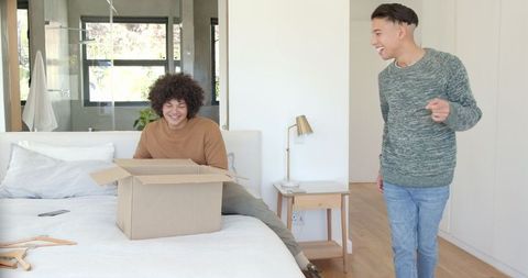 Friends Unpacking Moving Box Together on Modern Bedroom Bed