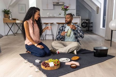 Couple sharing charcuterie and pouring wine during cozy indoor picnic on knit blanket