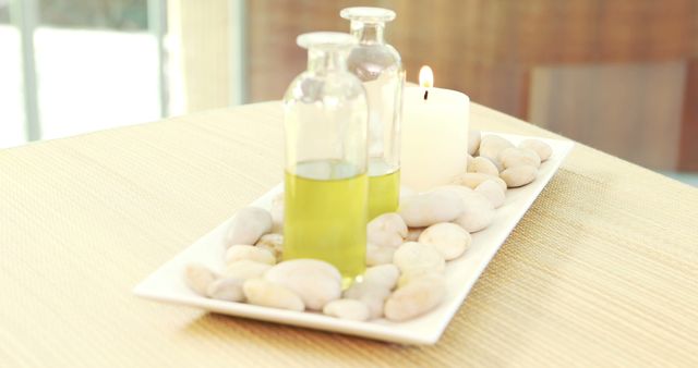 Two glass bottles containing yellow aromatherapy oils are placed on a white rectangular tray filled with white decorative stones alongside a lit white candle. This is ideal for use in blogs or advertising about wellness, spa treatments, massage therapy, relaxation techniques, and tranquility retreat promotions.