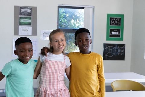 Diverse elementary students linking arms, laughing and smiling in bright classroom