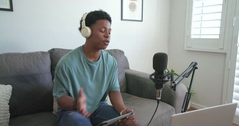 African american man recording podcast on couch wearing headphones with mic and laptop
