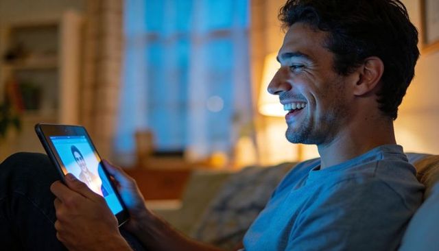 Smiling man using tablet for video call on sofa during cozy evening at home with warm lamp