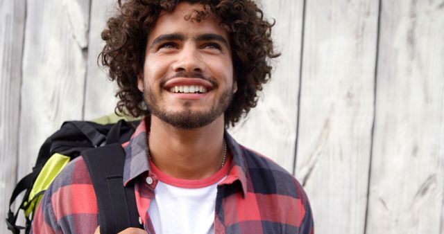 Happy biracial man with curly hair standing and smiling by wooden wall on sunny day, copy space. City break, summer, travel, vacations and lifestyle, unaltered.