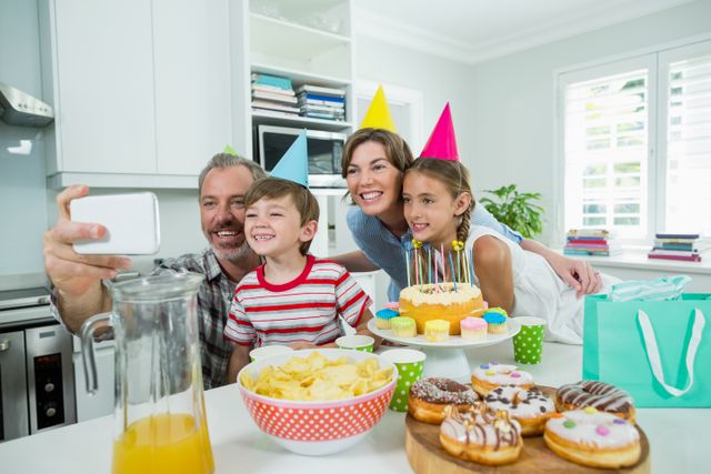 Family celebrating a birthday in their kitchen, taking a selfie to capture the moment. Parents and children are wearing party hats and smiling. The table is filled with a birthday cake, donuts, chips, and juice. Ideal for use in family-oriented advertisements, birthday party promotions, and lifestyle blogs.