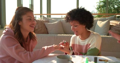 Friends enjoying at-home manicure, laughing while painting nails on sunlit living room sofa