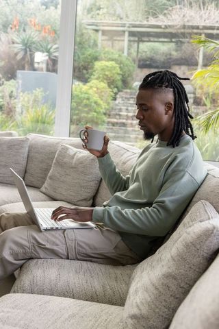 African american man using laptop and holding coffee mug on cozy sofa with garden view