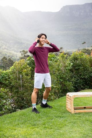 Young asian man enjoying music outdoors with headphones on manicured lawn by mountain ridge