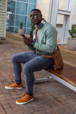 African american man sitting on bench holding smartphone and crossbody bag, urban courtyard