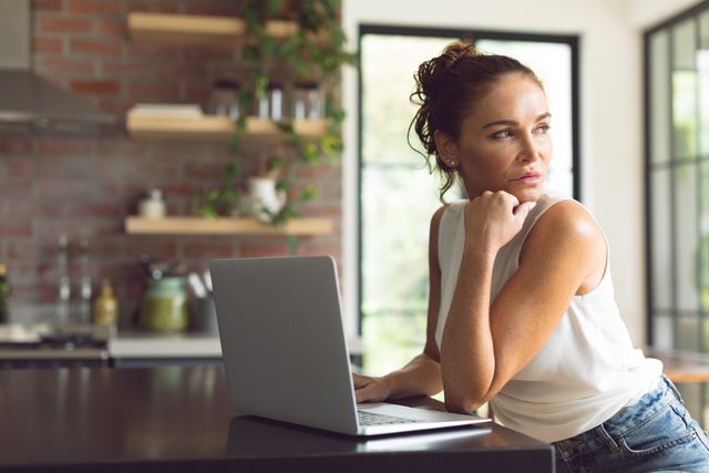 Beautiful woman looking away while using laptop on worktop in kitchen at comfortable home 
