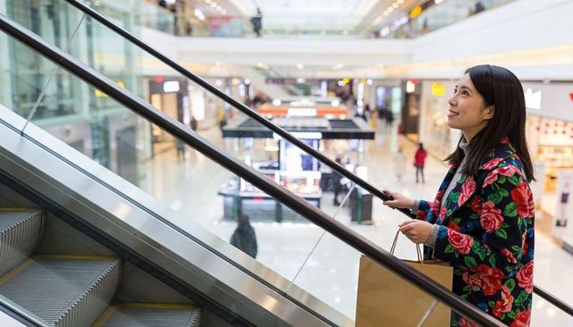 Woman riding escalator in modern mall wearing floral coat holding shopping bag smiling