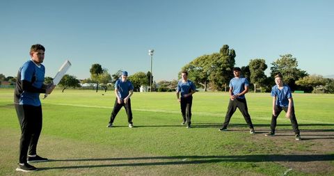 Batter swinging cricket bat while teammates readying to field on sunlit green ground