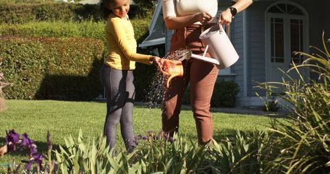 Mom and daughter watering flower bed with pink and orange watering cans in sunlit front yard
