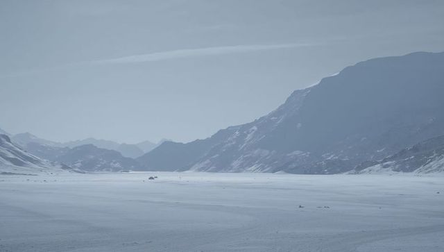 Vast frozen alpine valley with distant vehicle cluster, expedition tents and snow tracks