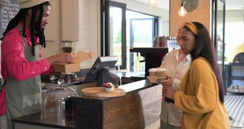 Barista packing pastry box while diverse customers picking up coffee at cozy cafe counter