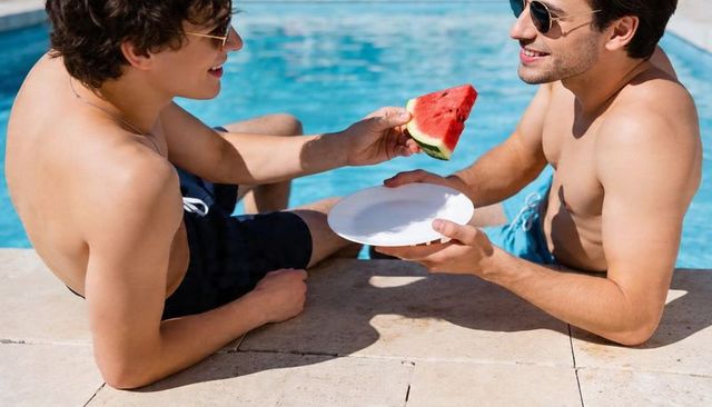 Two men sharing watermelon poolside wearing sunglasses and swim trunks summer relaxation