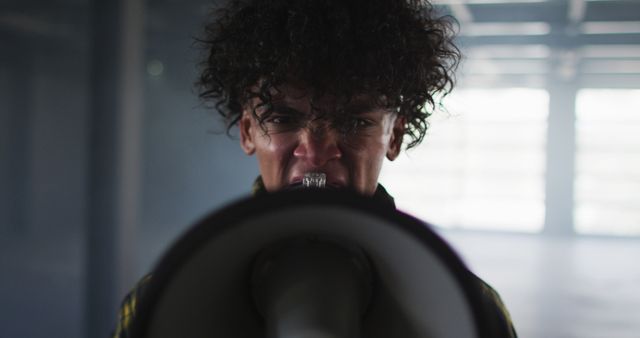 Close-up of individual with curly hair shouting into a megaphone in an industrial warehouse. Can be used for themes of activism, protest, conveying emotions such as anger and determination, leadership roles, or portraying heated speeches and calls to action.