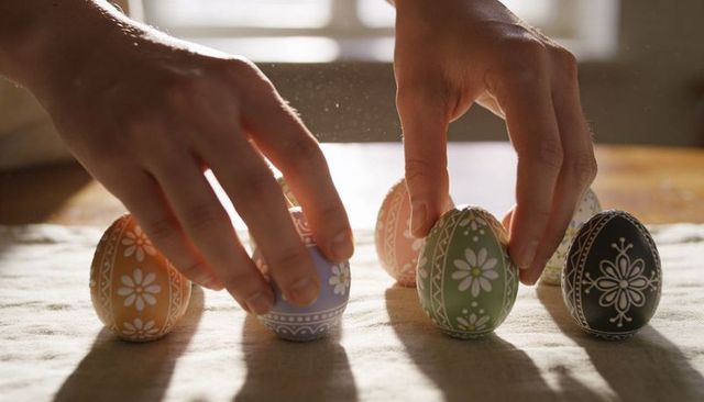 Sunlit hands placing hand-painted easter eggs with floral folk patterns on rustic table
