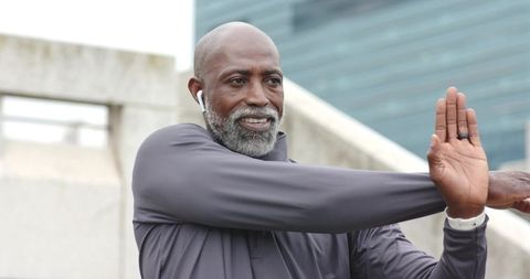 Senior African American man stretching chest on city stairs wearing earbuds, athletic top