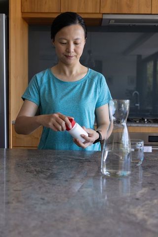 Asian woman opening medicine container at modern kitchen island with glass carafe and teal tee