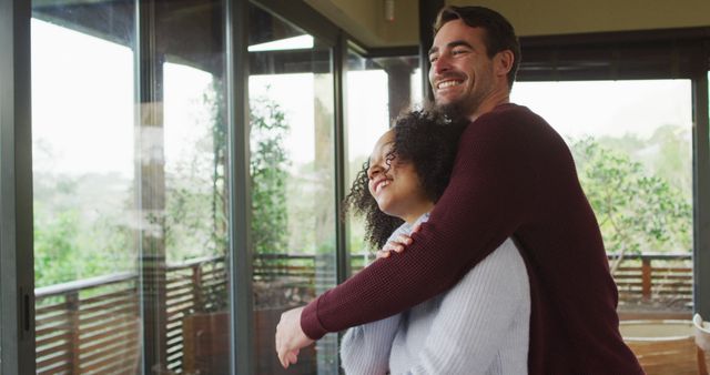 Happy diverse couple embracing and laughing in living room, looking out of window in the countryside. spending free time together at home.