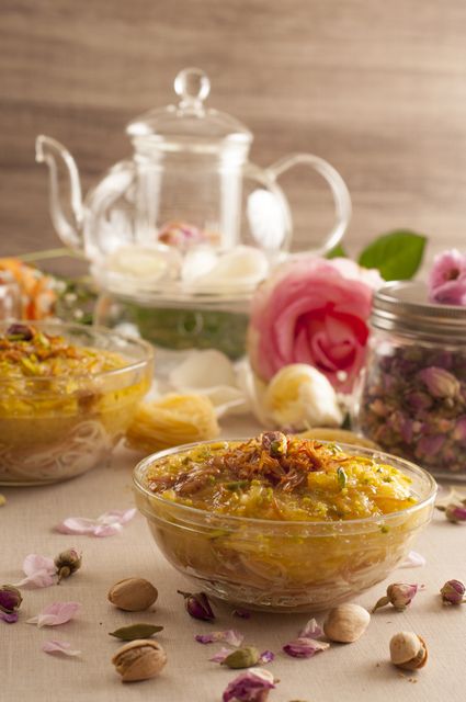 Traditional Persian saffron dessert served in glass bowls, garnished with pistachios and dried flowers, paired with a transparent tea pot. Roses and petals scattered around create a fragrant and inviting setting. Ideal for illustrating Persian cuisine, traditional celebrations, or Middle Eastern culinary articles.