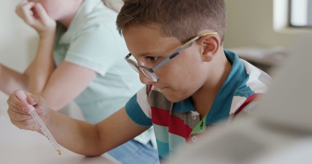 Boy wearing glasses is concentrating on a science experiment in a classroom setting, holding a tube and looking closely at it. Ideal for use in educational materials, school websites, or advertising for educational products.