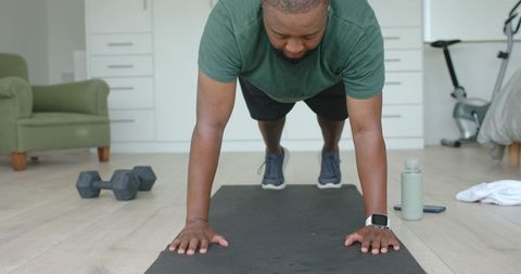 African american man performing plank at home workout with smartwatch and dumbbell nearby