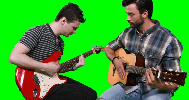 Two men playing guitar, one with an acoustic and the other with an electric, against a green screen background. They are focused on their instruments, suggesting a sense of concentration and collaboration. This can be used for music tutorials, advertisements for music lessons, or promotional materials for bands and live music events.