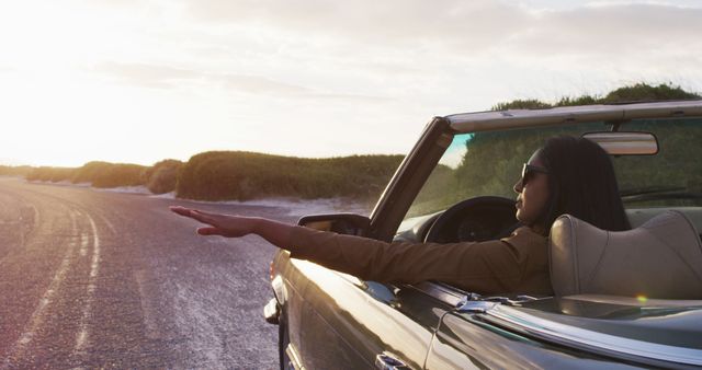 Young woman driving convertible on open road at sunset, arm resting out window. Perfect for themes of travel, freedom, leisure, summer vacations, and road trips. Ideal for lifestyle blogs, advertisements, tourism campaigns, and automotive promotions.