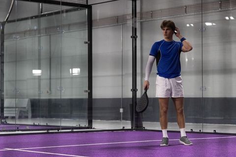 Padel player standing on purple court by glass wall preparing serve and looking focused