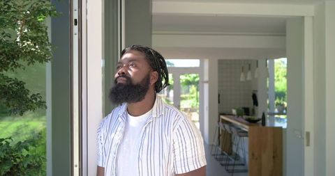 African american man standing in sliding glass doorway gazing at garden at modern home
