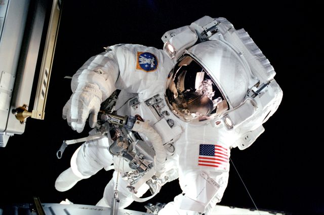 An astronaut in a white spacesuit conducting a spacewalk outside the International Space Station (ISS). The astronaut is preparing to work with the Materials International Space Station Experiment (MISSE), collecting data on how materials interact with the space environment. Useful for topics on space exploration, scientific research, NASA missions, and technology advancements in space.