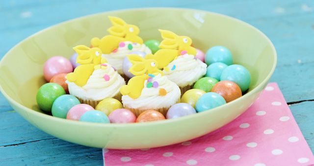 A bowl of colorful Easter-themed cupcakes and candies, with pastel colors and bunny decorations, sits on a blue wooden surface with copy space. Festive treats like these are often used to celebrate the spring season and Easter holiday.